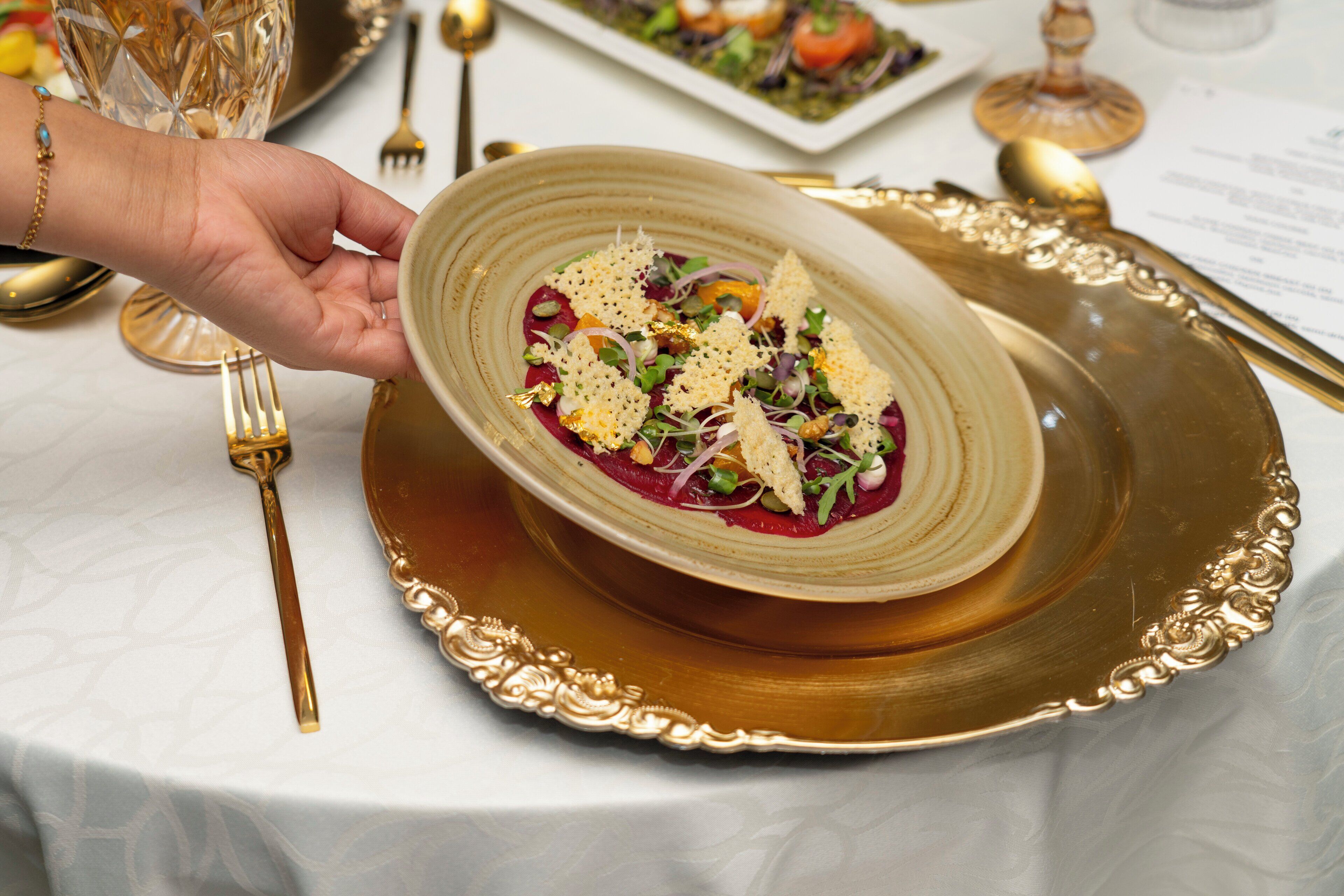 A hand holding a plate with a dish, placed on a gold-rimmed plate on a table with a white tablecloth.