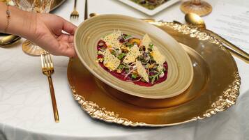 A hand holding a plate with a dish, placed on a gold-rimmed plate on a table with a white tablecloth.