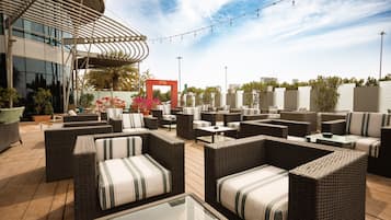 An outdoor seating area with wicker furniture and tables, a clear sky, and a building with a distinctive curved roof in the background.
