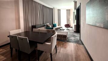 Una habitación de hotel moderna con zona de comedor, un conjunto de sofás, un sillón rojo y un cuadro grande en la pared.