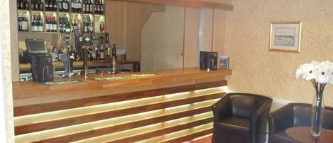 Una zona de bar con mostrador de madera, estantes llenos de botellas y un espacio de estar con dos sillas y una mesa.