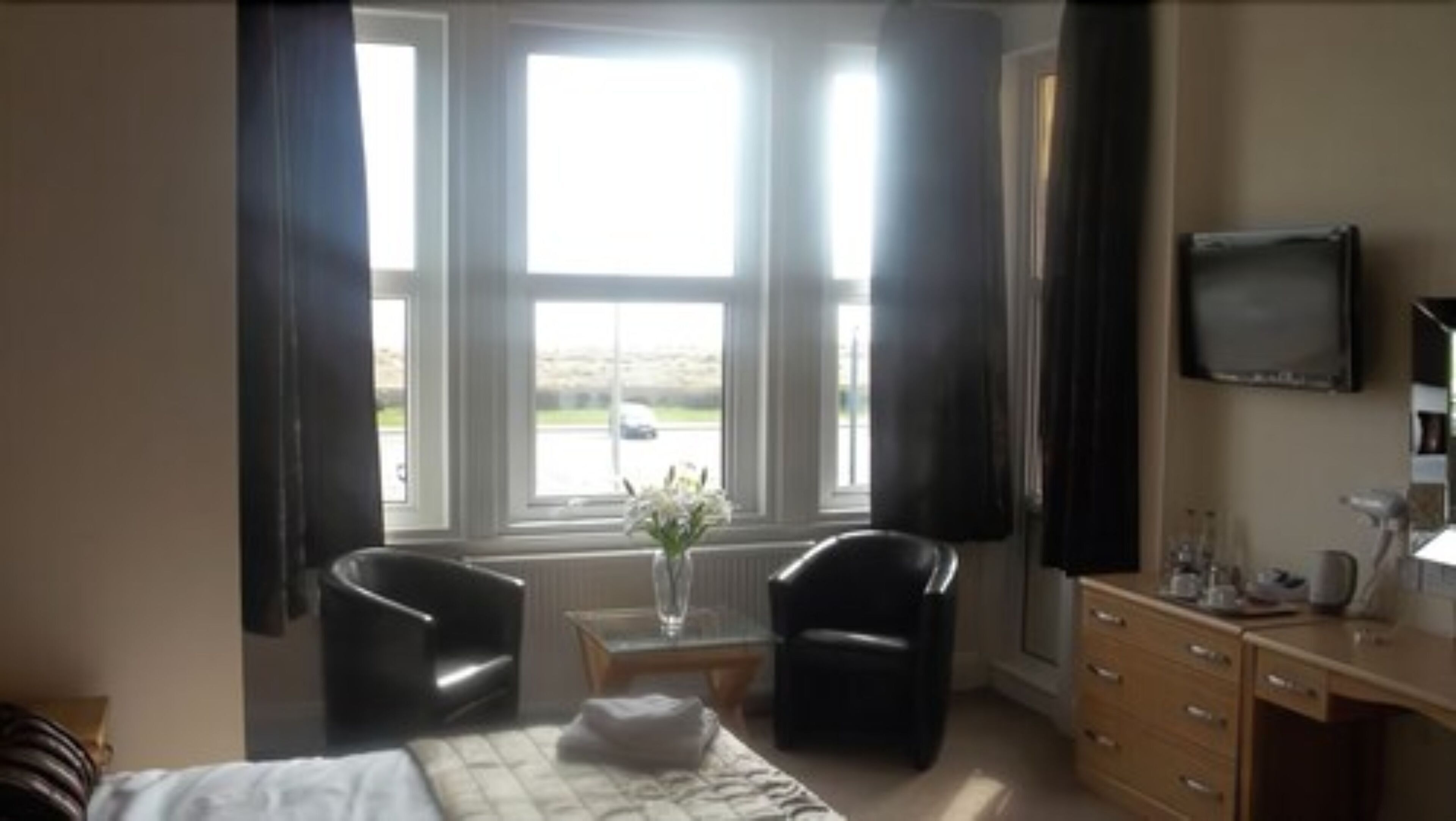 A hotel room with a bed, two armchairs, a small table with a vase, a dresser, and a TV on the wall.