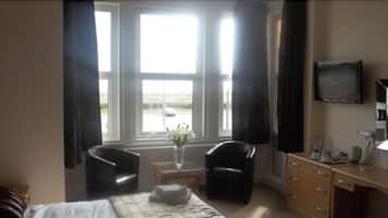 A hotel room with a bed, two armchairs, a small table with a vase, a dresser, and a TV on the wall.