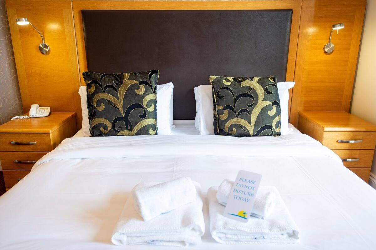 Ein Hotelzimmer mit einem großen Bett, zwei Zierkissen, einem Nachttisch mit Telefon und einem Schild am Bett.