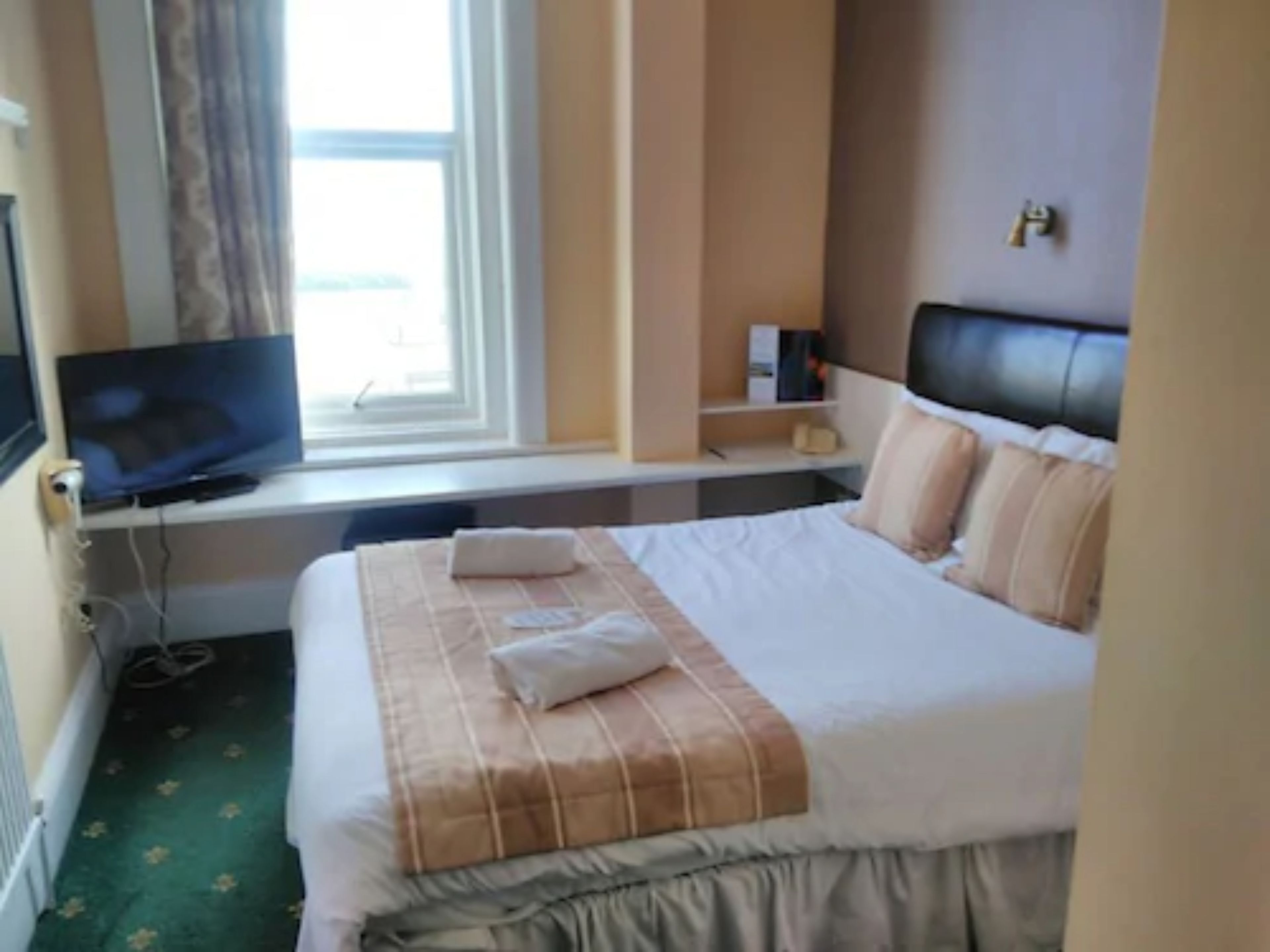 A hotel room with a bed, a television on a desk, a window with curtains, and a bedside table with a lamp.