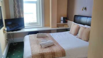 A hotel room with a bed, a television on a desk, a window with curtains, and a bedside table with a lamp.
