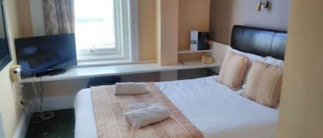 Une chambre d’hôtel avec un lit, un téléviseur posé sur un bureau, une fenêtre avec des rideaux et une table de chevet avec une lampe.