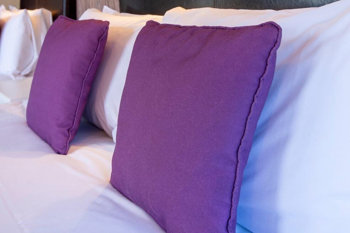 Habitación de hotel con pared de madera texturizada, cabecero negro y ropa de cama blanca con almohadas moradas.