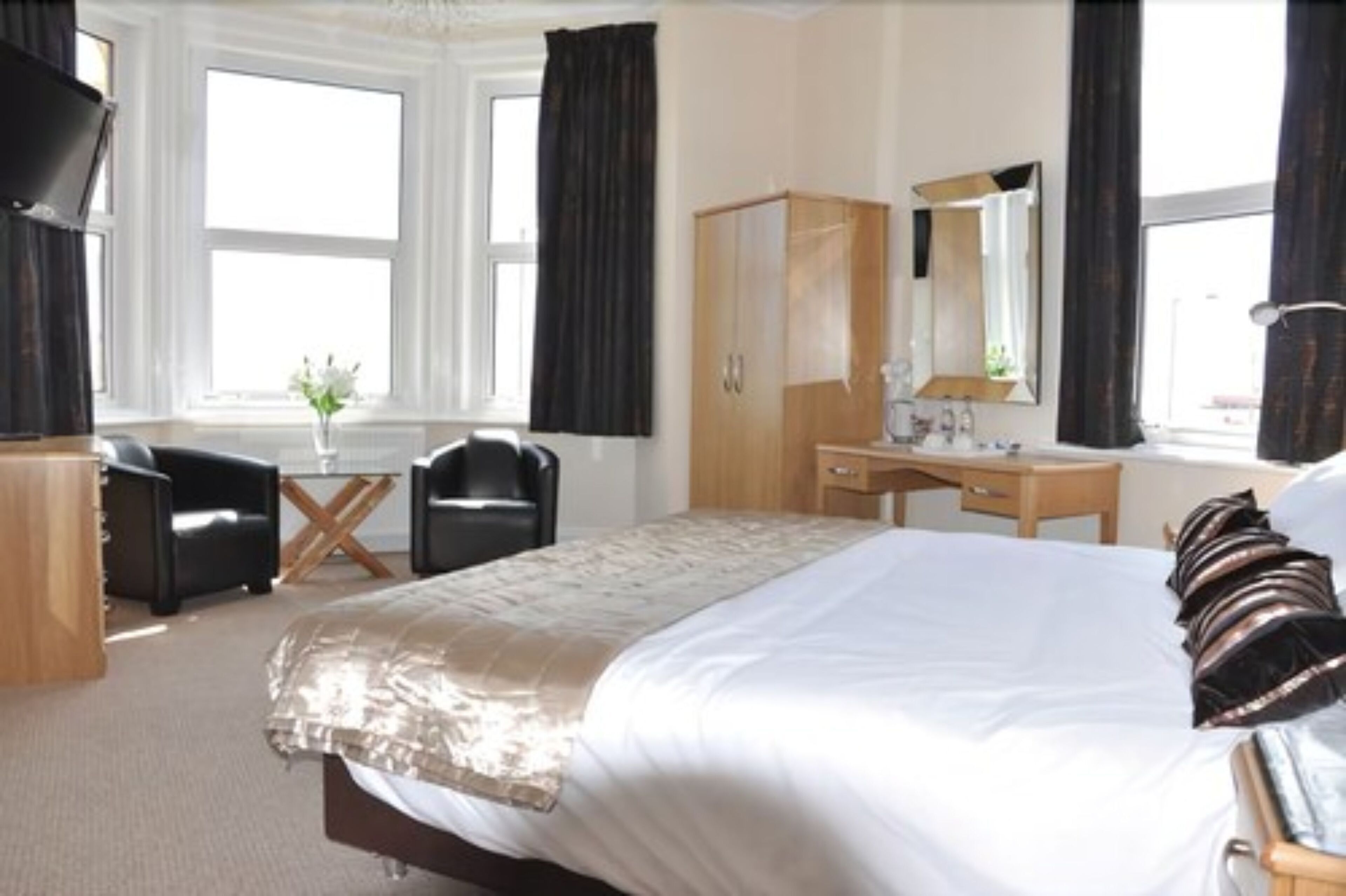 A hotel room with a large bed, a wooden wardrobe, a desk with a chair, a mirror, and a window with curtains.