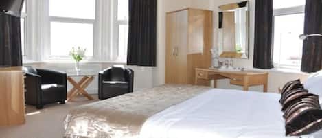 A hotel room with a large bed, a wooden wardrobe, a desk with a chair, a mirror, and a window with curtains.