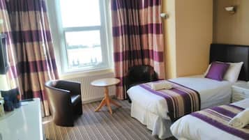 A hotel room with two beds, a small round table, a chair, and a view of the sea through a window.