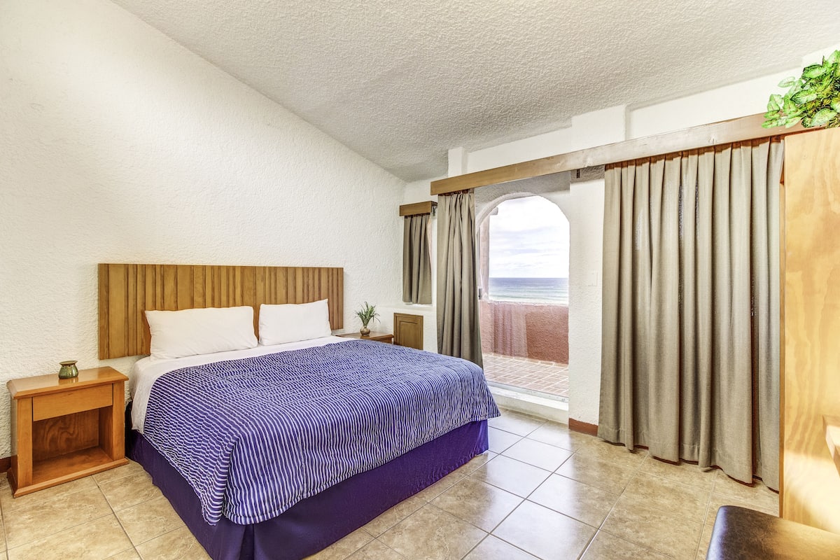 A bedroom with a bed, wooden headboard, nightstands, a potted plant, and a view of the beach through large windows.