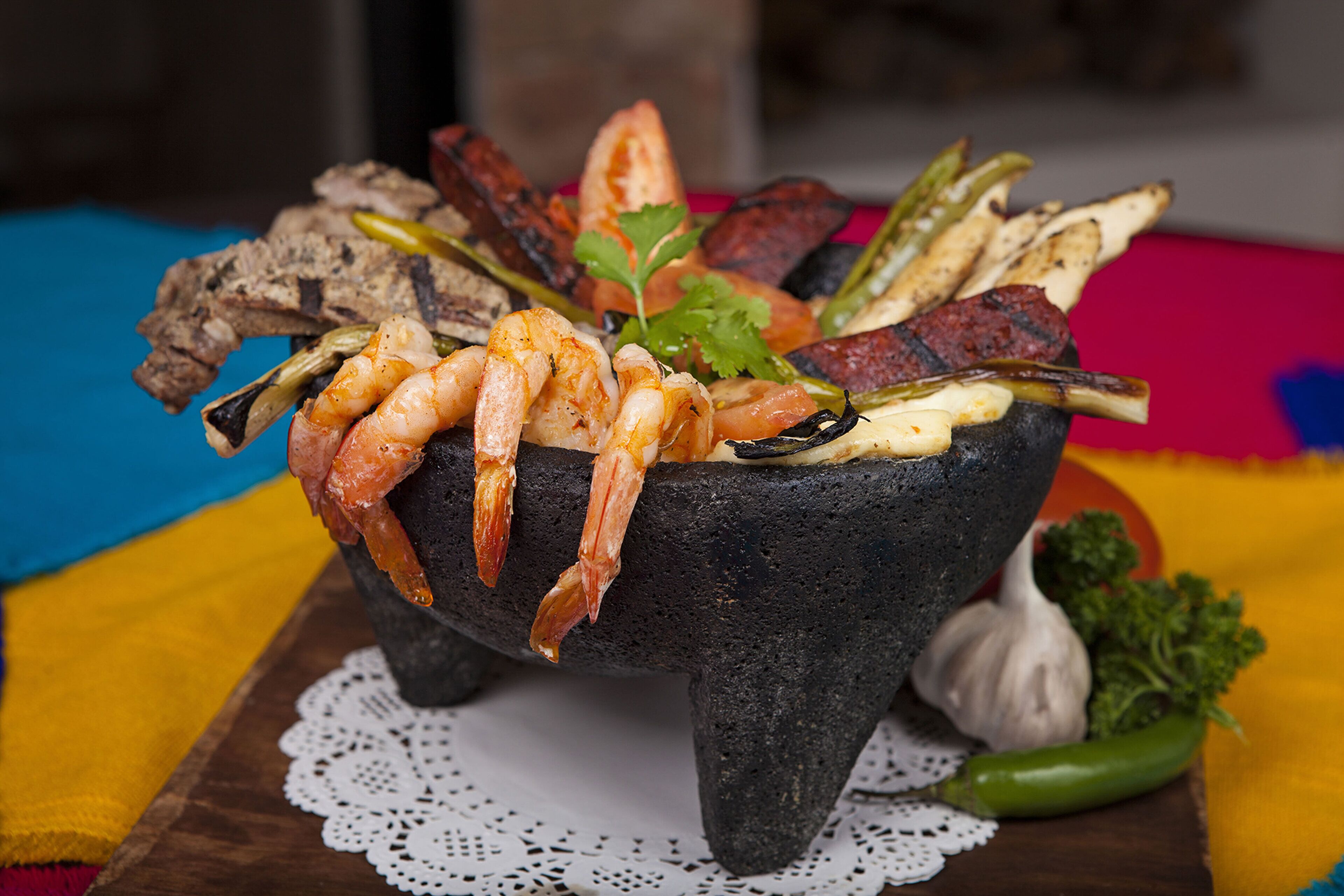 A stone bowl filled with grilled shrimp, vegetables, and meat, placed on a wooden table with a colorful tablecloth.