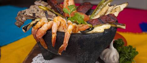 A stone bowl filled with grilled shrimp, vegetables, and meat, placed on a wooden table with a colorful tablecloth.