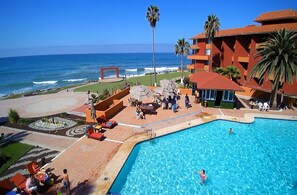 A hotel pool area with a view of the ocean, palm trees, and a red-roofed building. - Puerto Nuevo Baja Hotel & Villas (Playas de Rosarito)