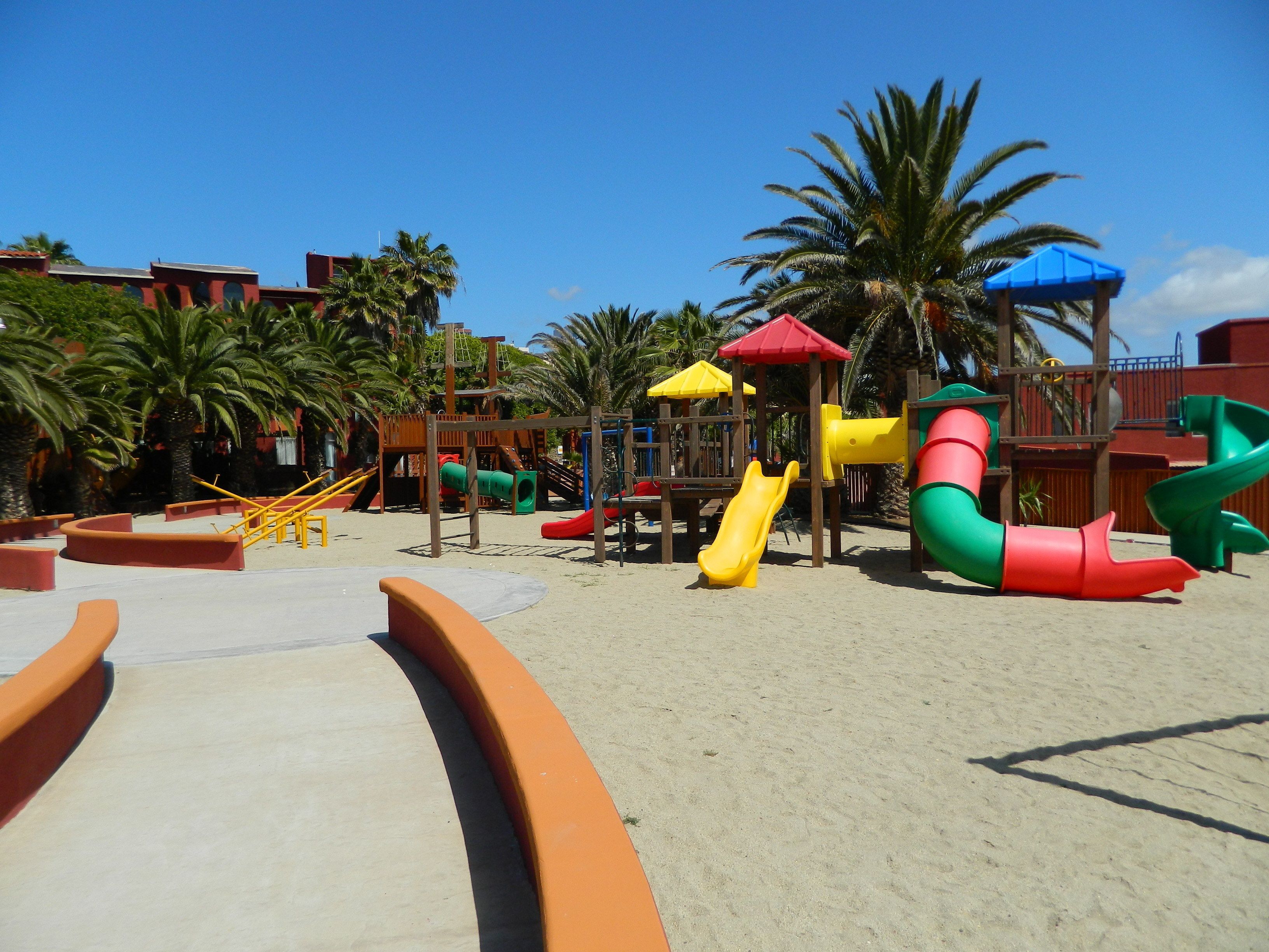A playground with slides and climbing structures, surrounded by palm trees and buildings.