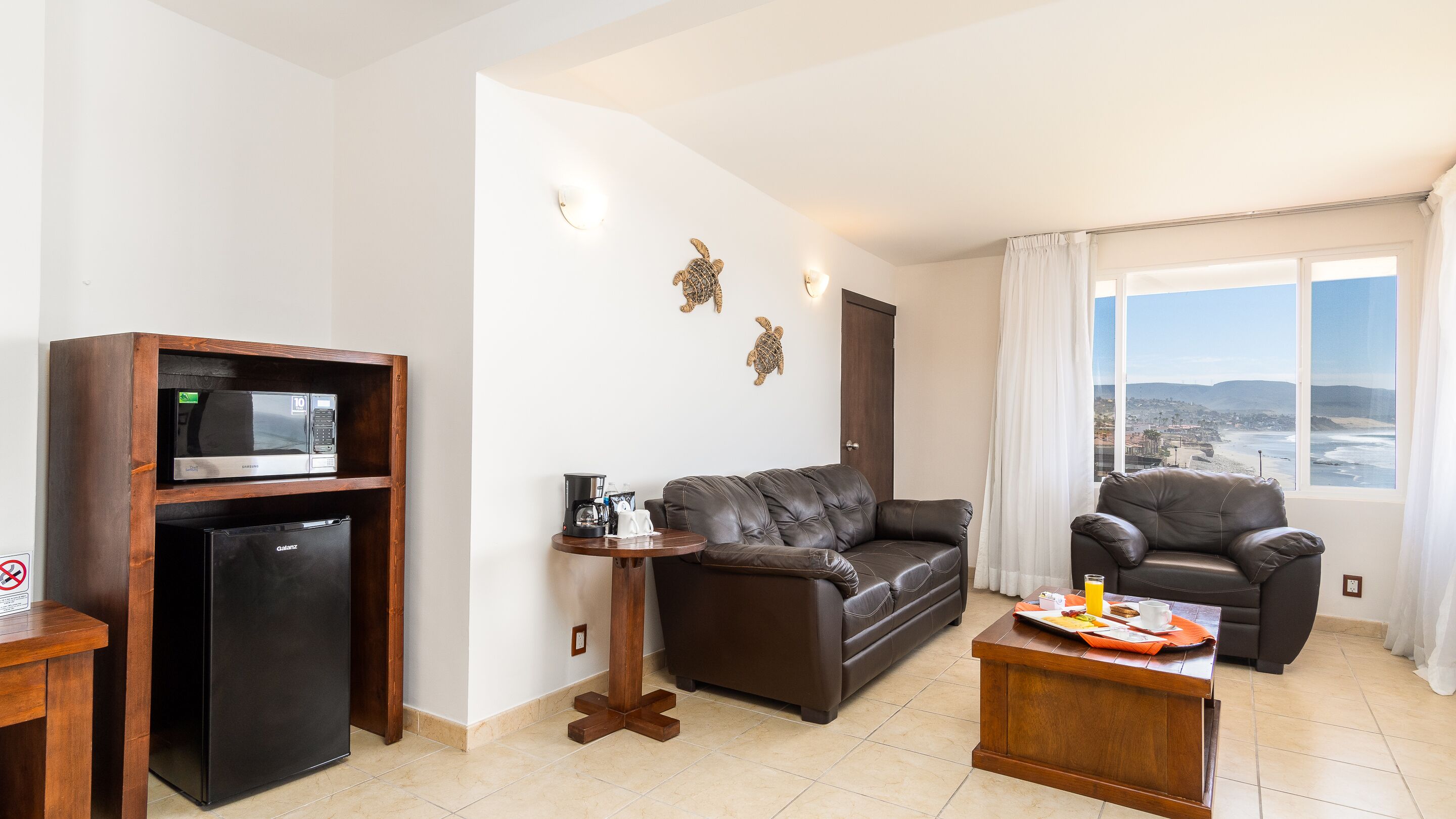 A living room with a sofa, two armchairs, a coffee table, a refrigerator, and a microwave. There is a window with a view of the beach and a door with a towel rack.