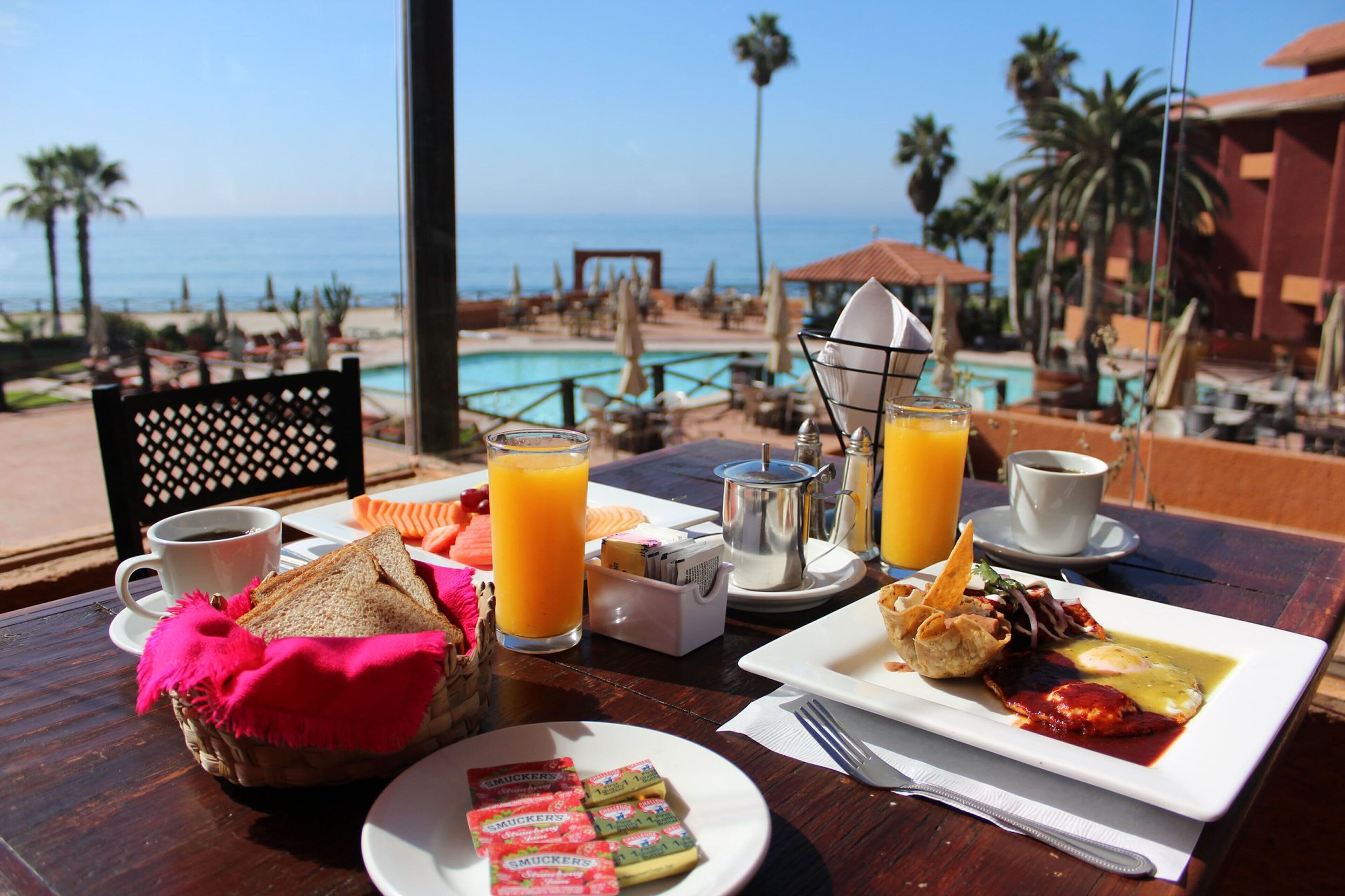 Une table de petit-déjeuner avec vue sur la plage et la piscine, comprenant des assiettes de nourriture, des boissons et un panier de pain.