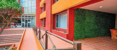 A building with a red exterior, a glass entrance, and a green wall. There are steps leading to the entrance and a railing in the foreground.