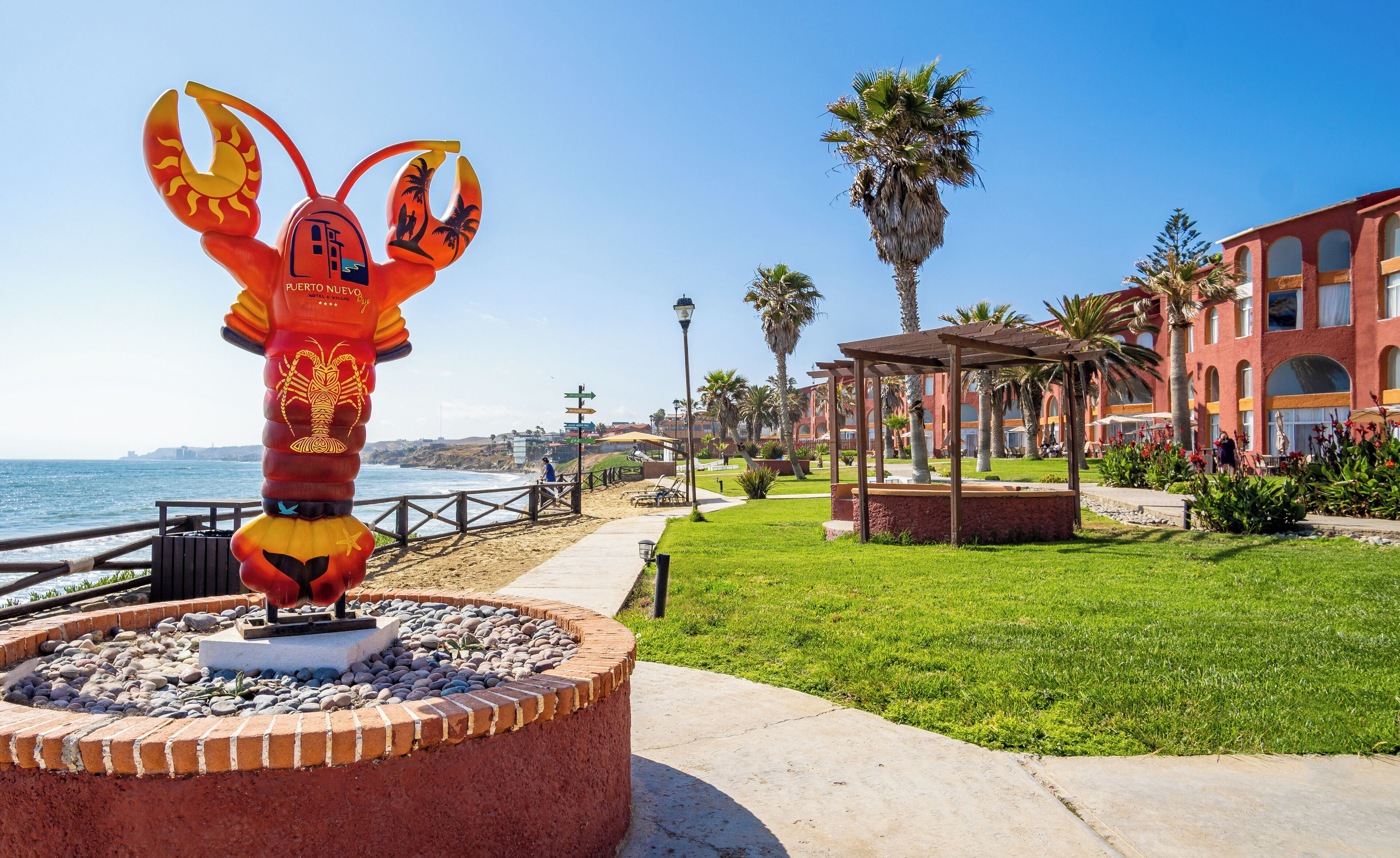 A coastal hotel with a prominent lobster sculpture, a beach promenade, and palm trees.