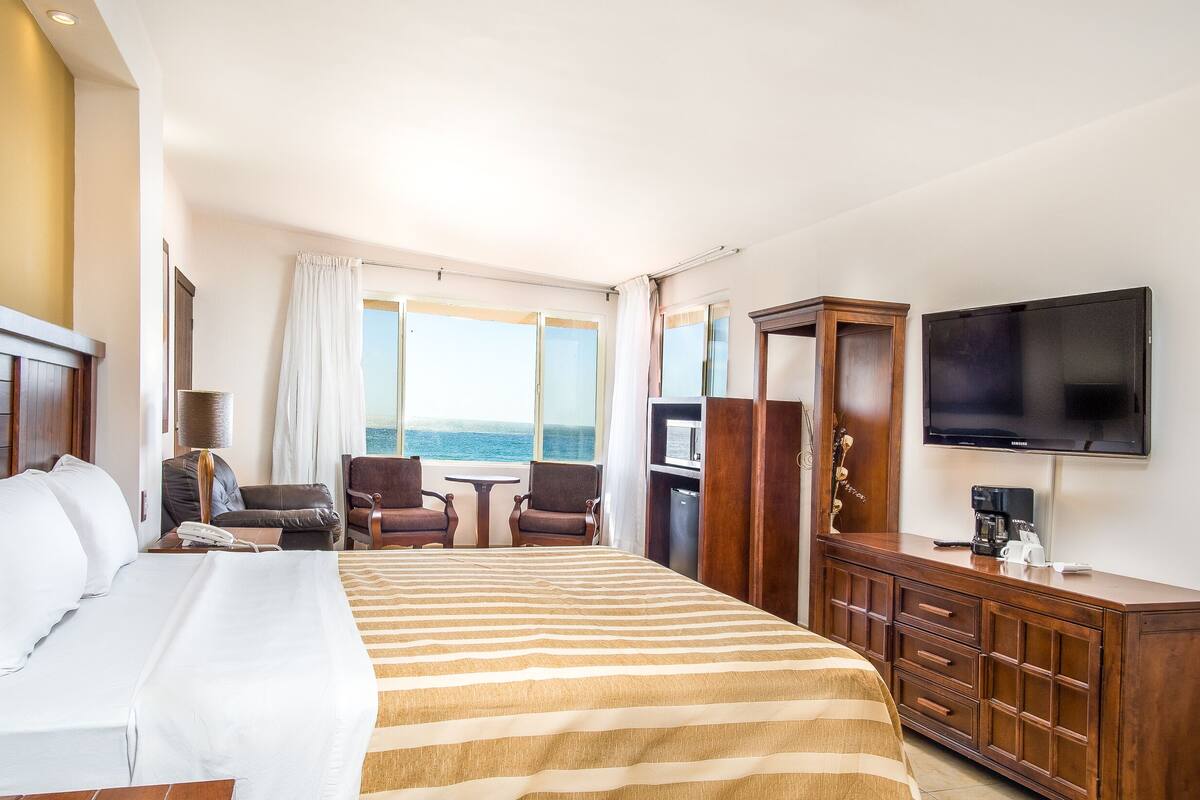 A hotel room with a large bed, a TV, a desk with a chair, and a view of the sea.