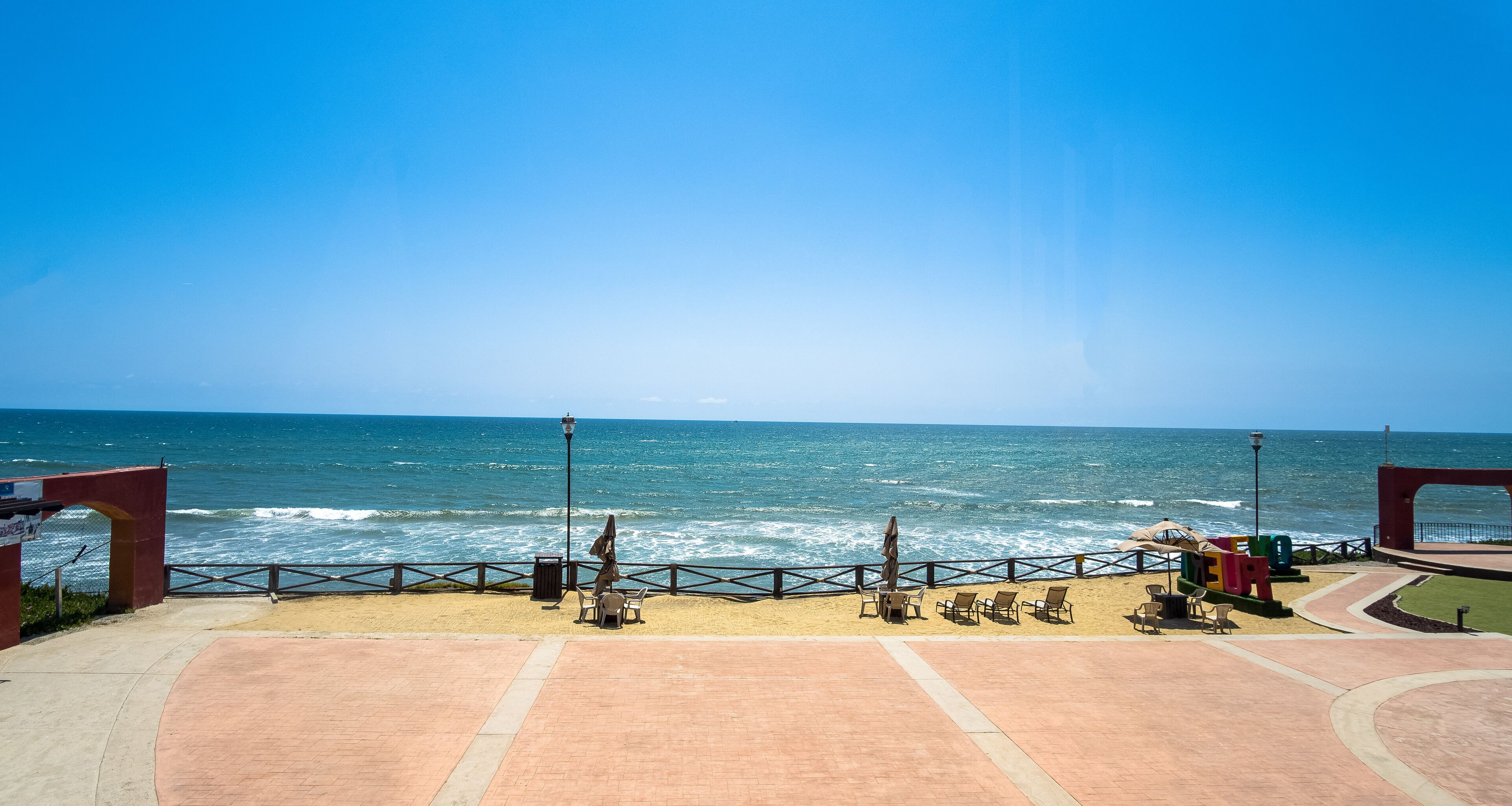 Un paseo marítimo en la playa con vista al océano, que incluye un sendero pavimentado, sillas de playa y sombrillas.