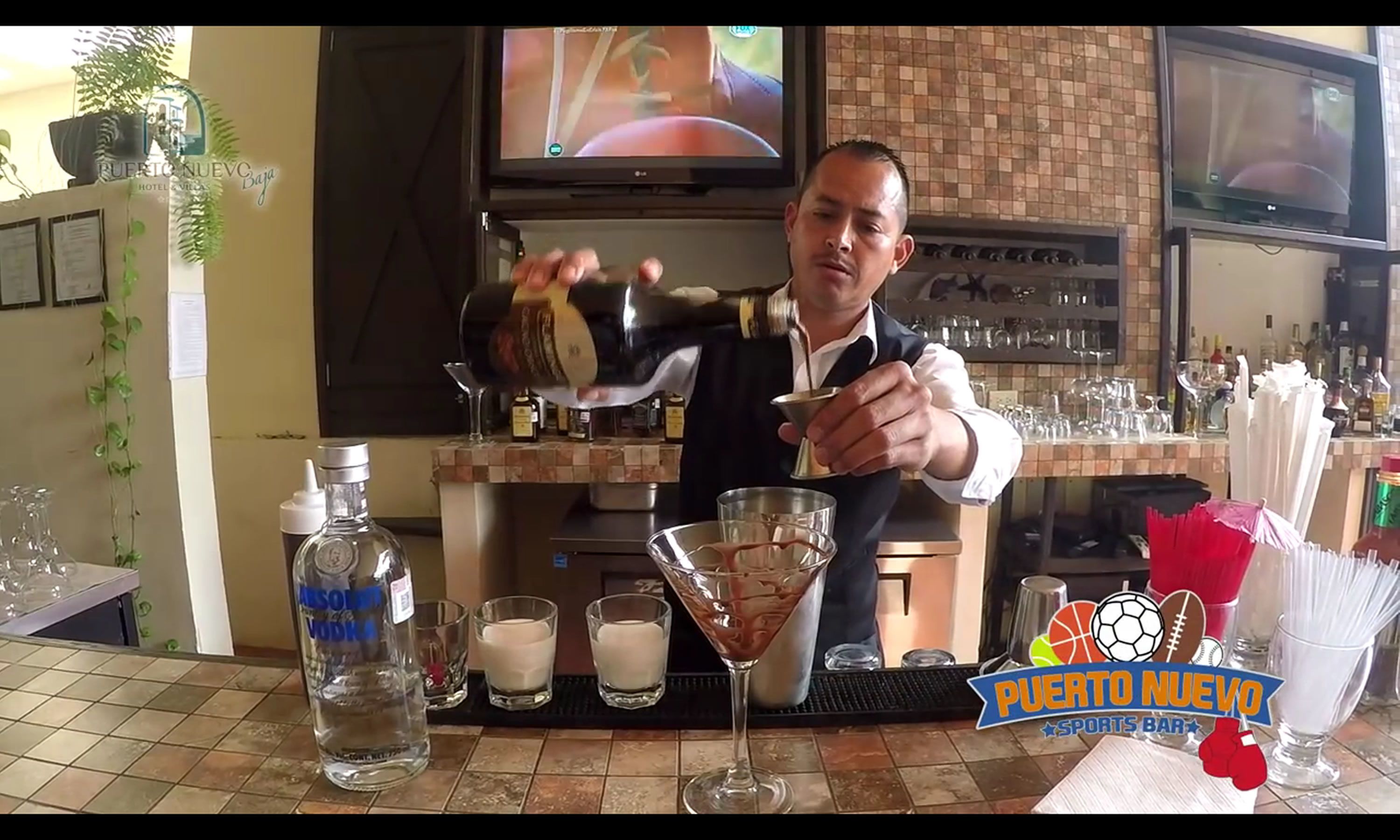 A bar area with a bartender preparing a drink, a bottle of Absolon Vodka, glasses, and a sports bar sign.