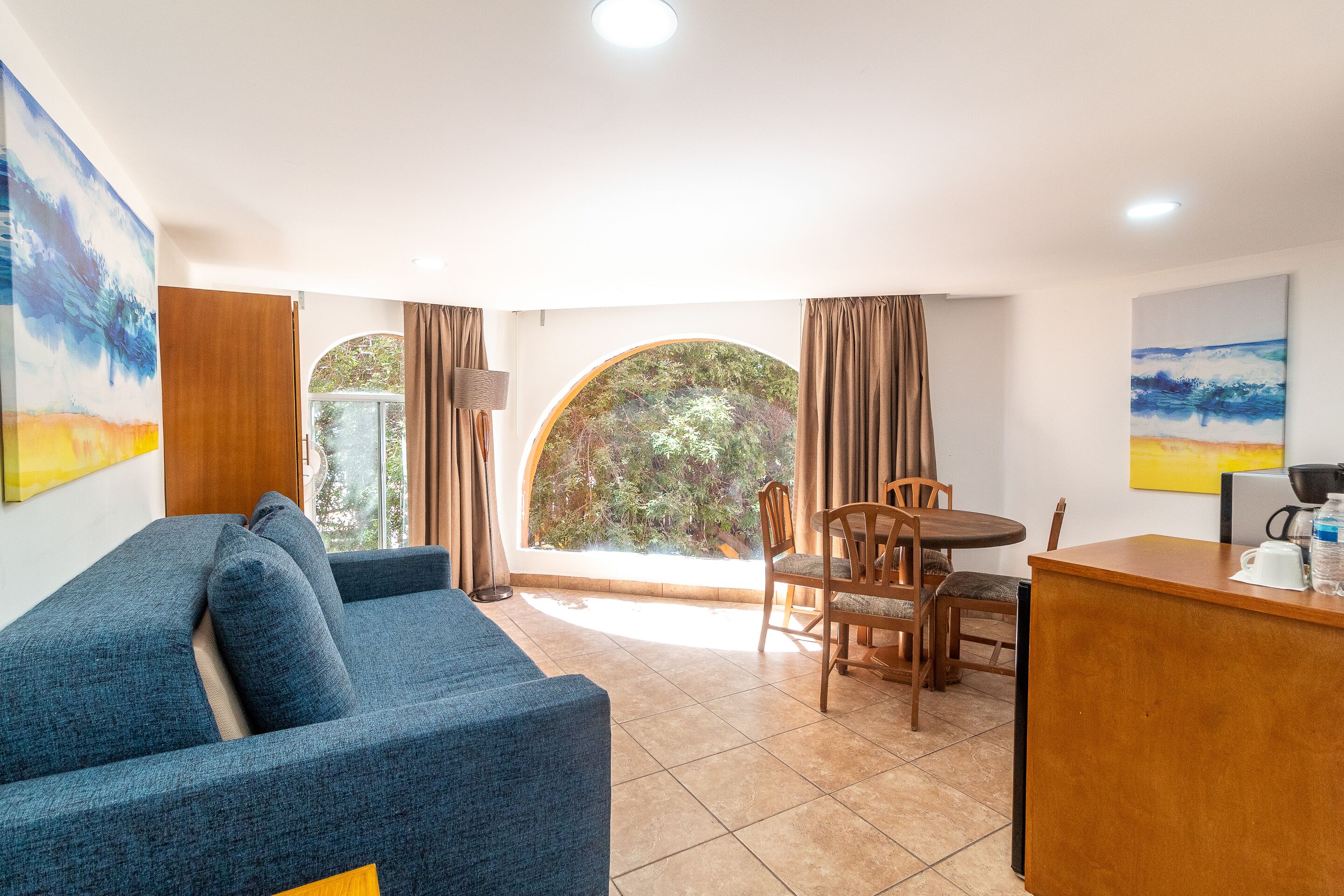 Un salón moderno con un sofá azul, una mesa de comedor de madera con sillas, una estación de café y ventanas grandes con cortinas.
