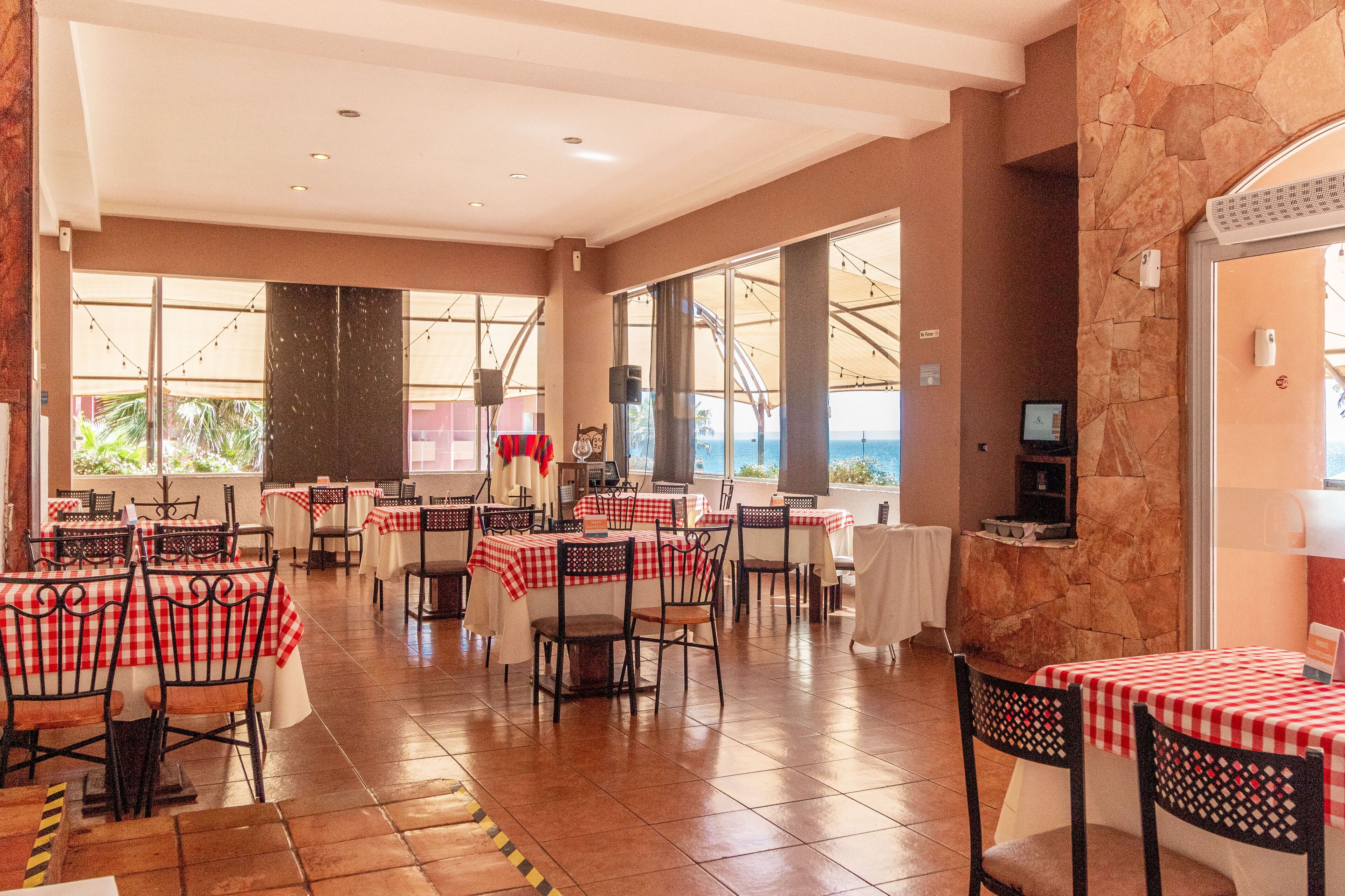 A spacious dining area with red and white checkered tablecloths, stone walls, and large windows offering a view of the ocean.