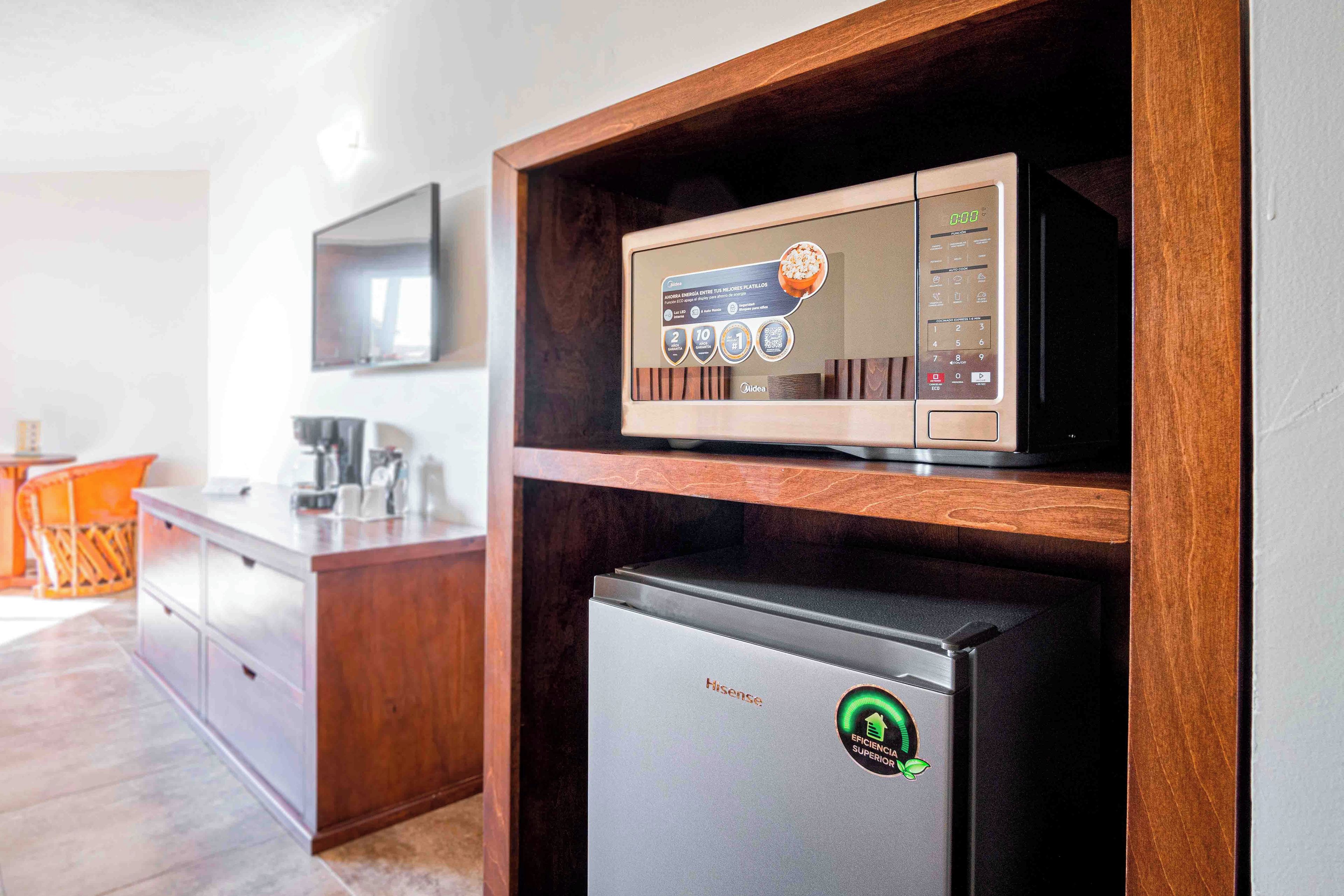 A compact kitchen with a microwave on a wooden shelf, a refrigerator below, and a TV mounted on the wall.