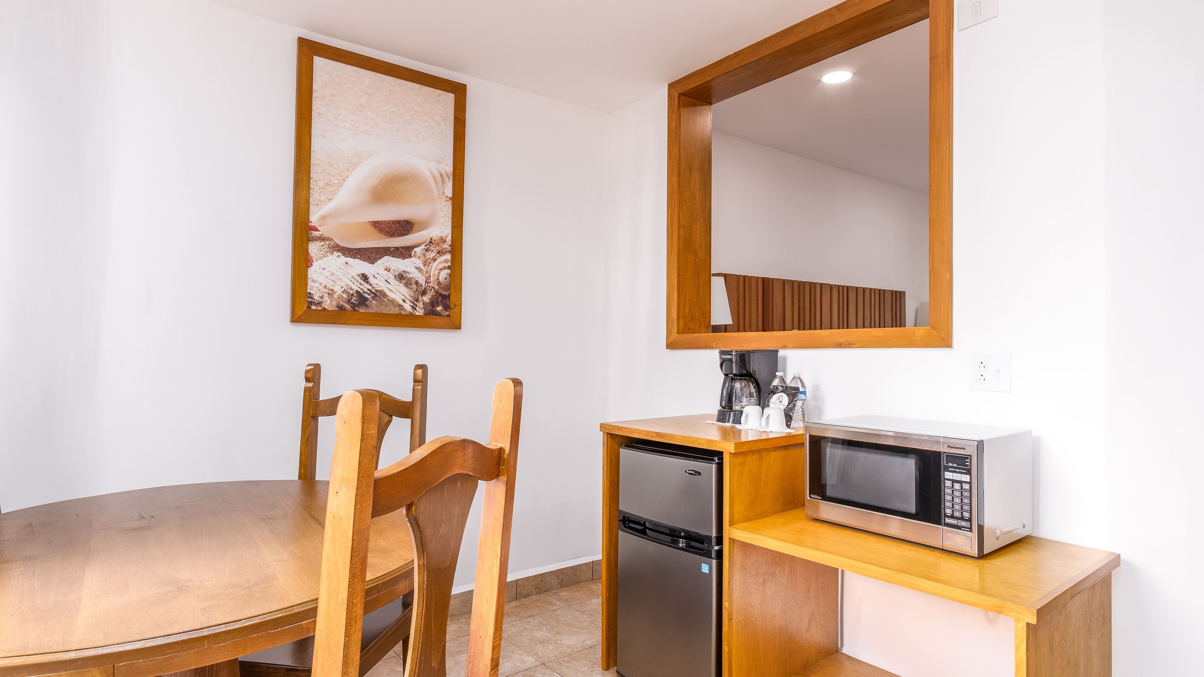 A compact kitchenette with a microwave, coffee maker, and small refrigerator. There is a wooden dining table and chairs, and a framed picture on the wall.