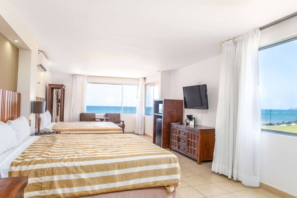 A hotel room with two beds, a TV, a desk, and a view of the sea.