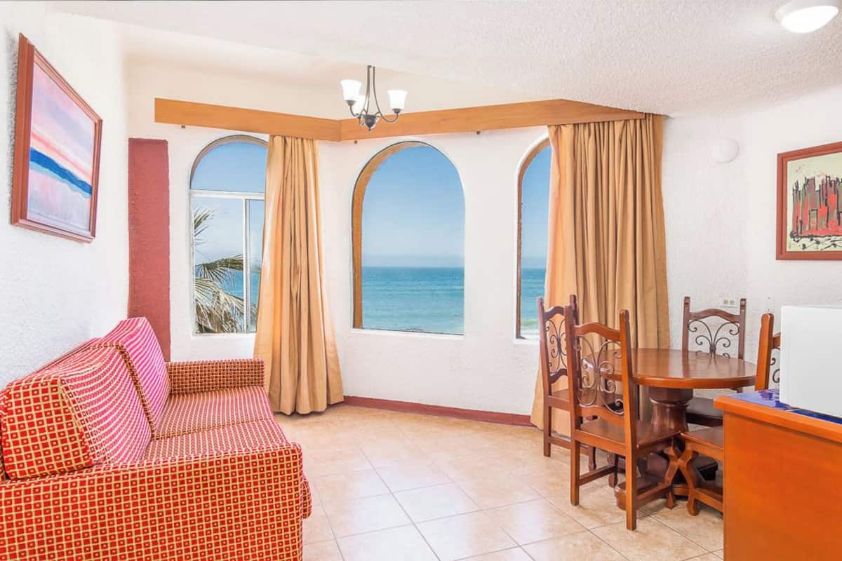 A room with a sofa, dining table, chairs, and a refrigerator. There are three windows with curtains, and a painting on the wall.