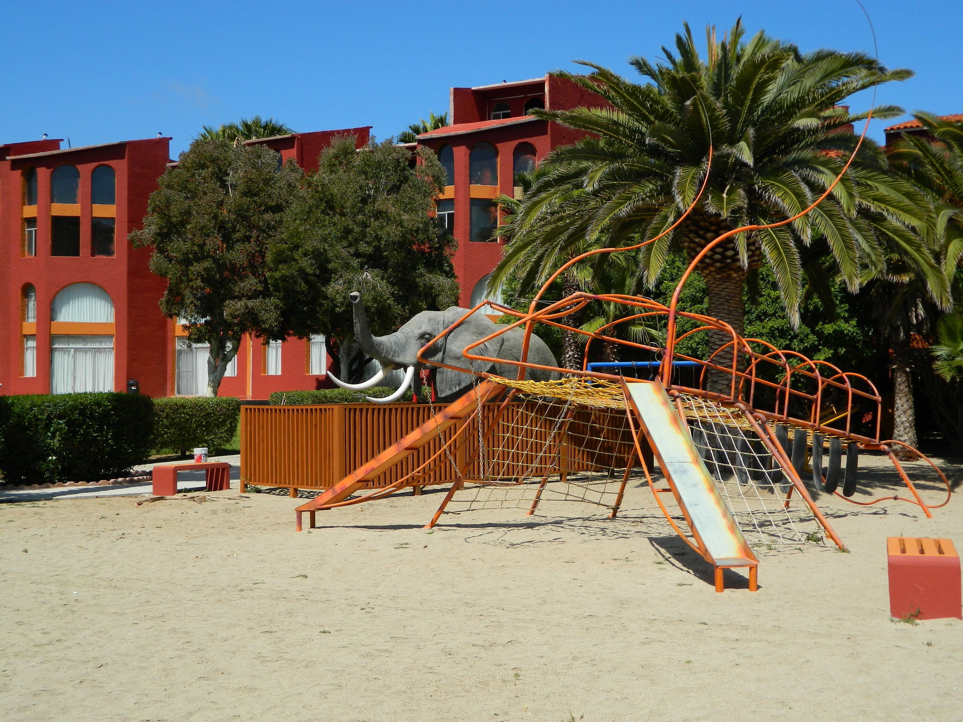 A playground with a slide and climbing structures, set against a red building with arched windows and palm trees.