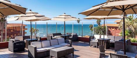 Une terrasse avec des espaces salons extérieurs, comprenant des canapés et des fauteuils, protégés par de grands parasols, et offrant une vue sur la mer.
