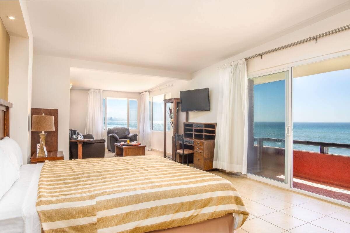 A hotel room with a large bed, a desk, a chair, a TV, and a balcony with a view of the sea.