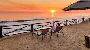 Vista a la playa con un atardecer, baranda de madera y dos sillas reclinables mirando al océano.