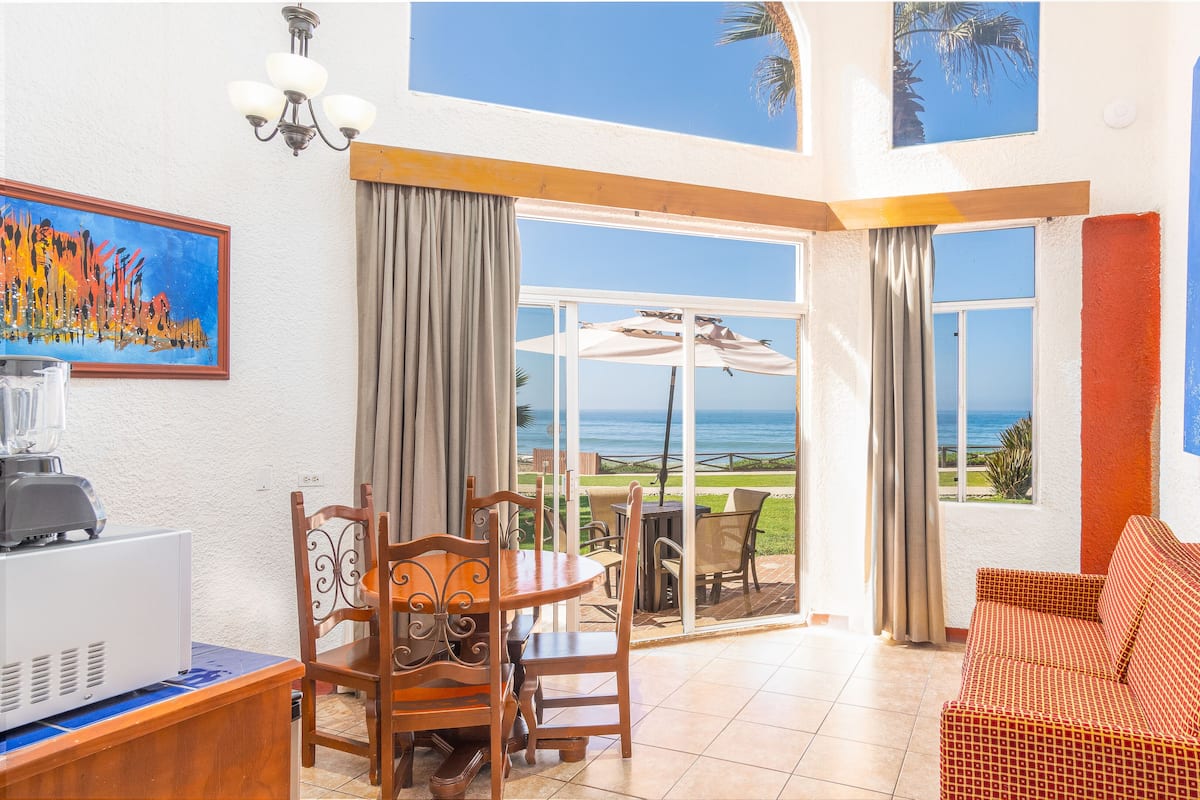 A room with a dining table, chairs, a sofa, a TV, and a view of the ocean.