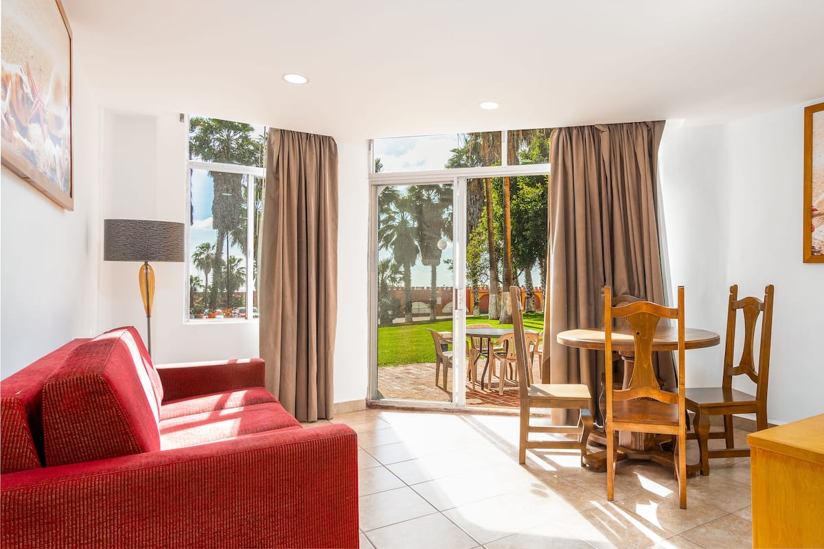 A hotel room with a red sofa, a dining table with chairs, a coffee maker, and a view of a garden through large windows.
