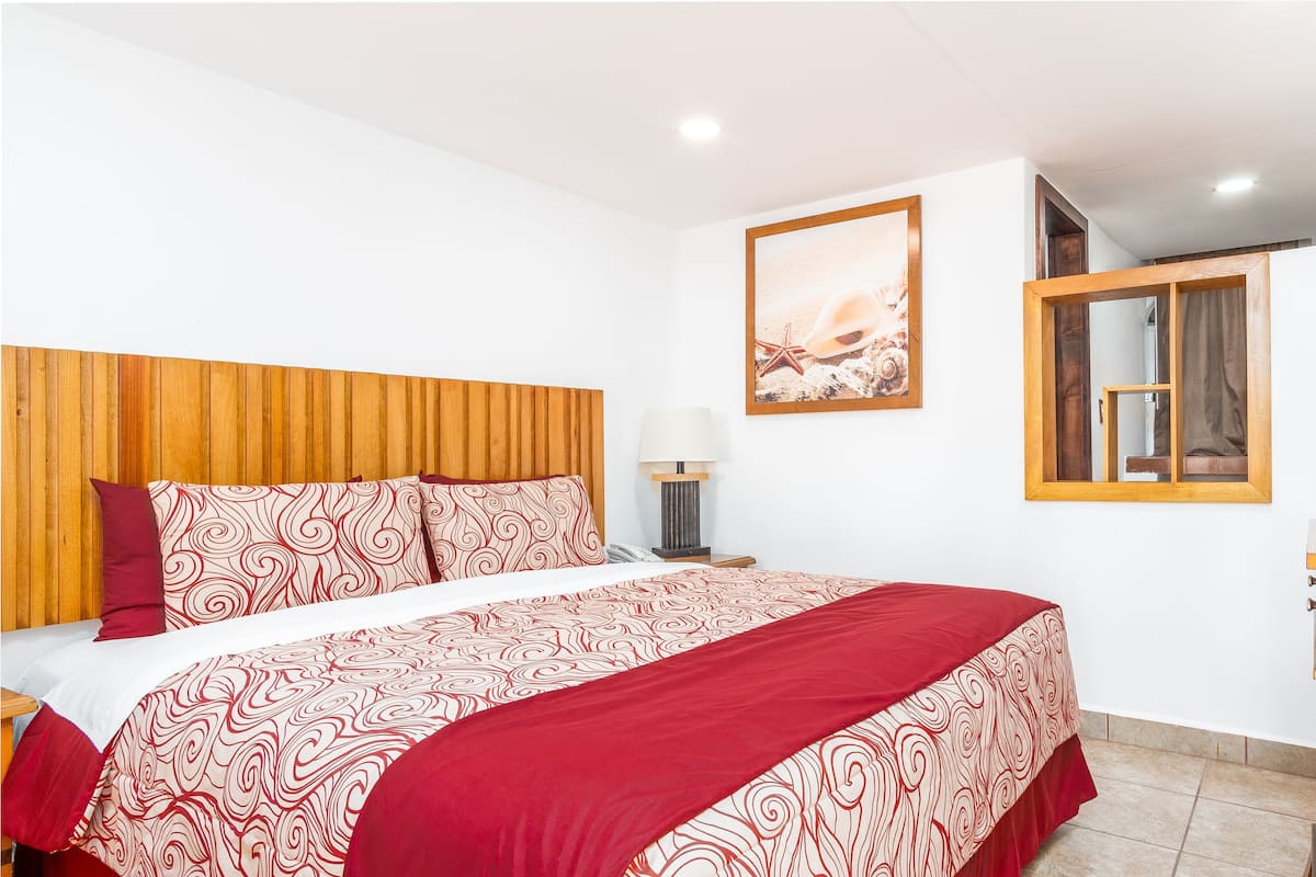 A hotel room with a wooden headboard, a bed with patterned bedding, a bedside table with a lamp, a TV, and a wooden dresser.