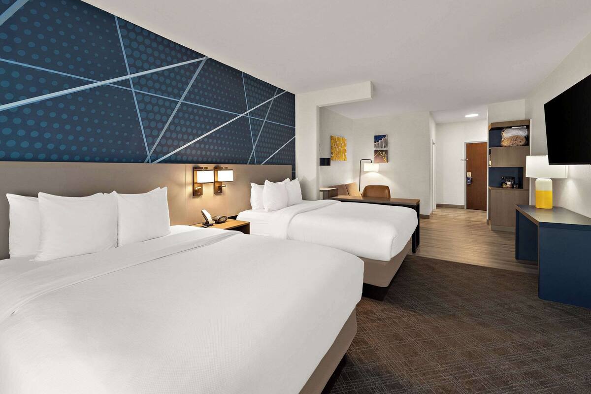 A hotel room with two beds, a desk, a chair, a TV, and a wall with a geometric pattern.