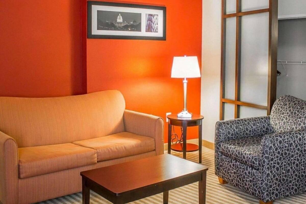 A hotel room with a sofa, a patterned armchair, a small table, a lamp, and a framed picture on the wall.