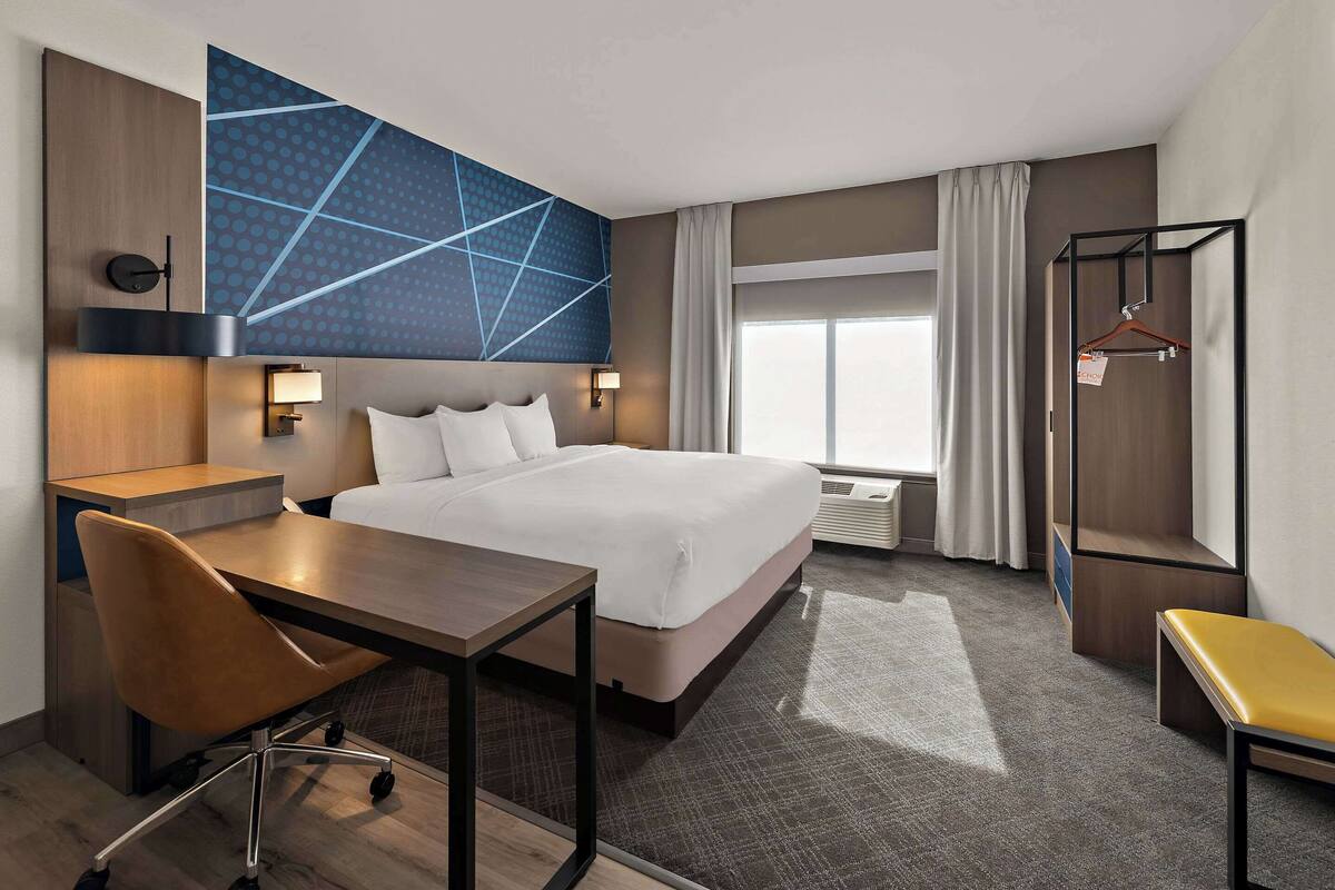A hotel room with a large bed, a desk, a chair, a hanging coat rack, and a large window with curtains.