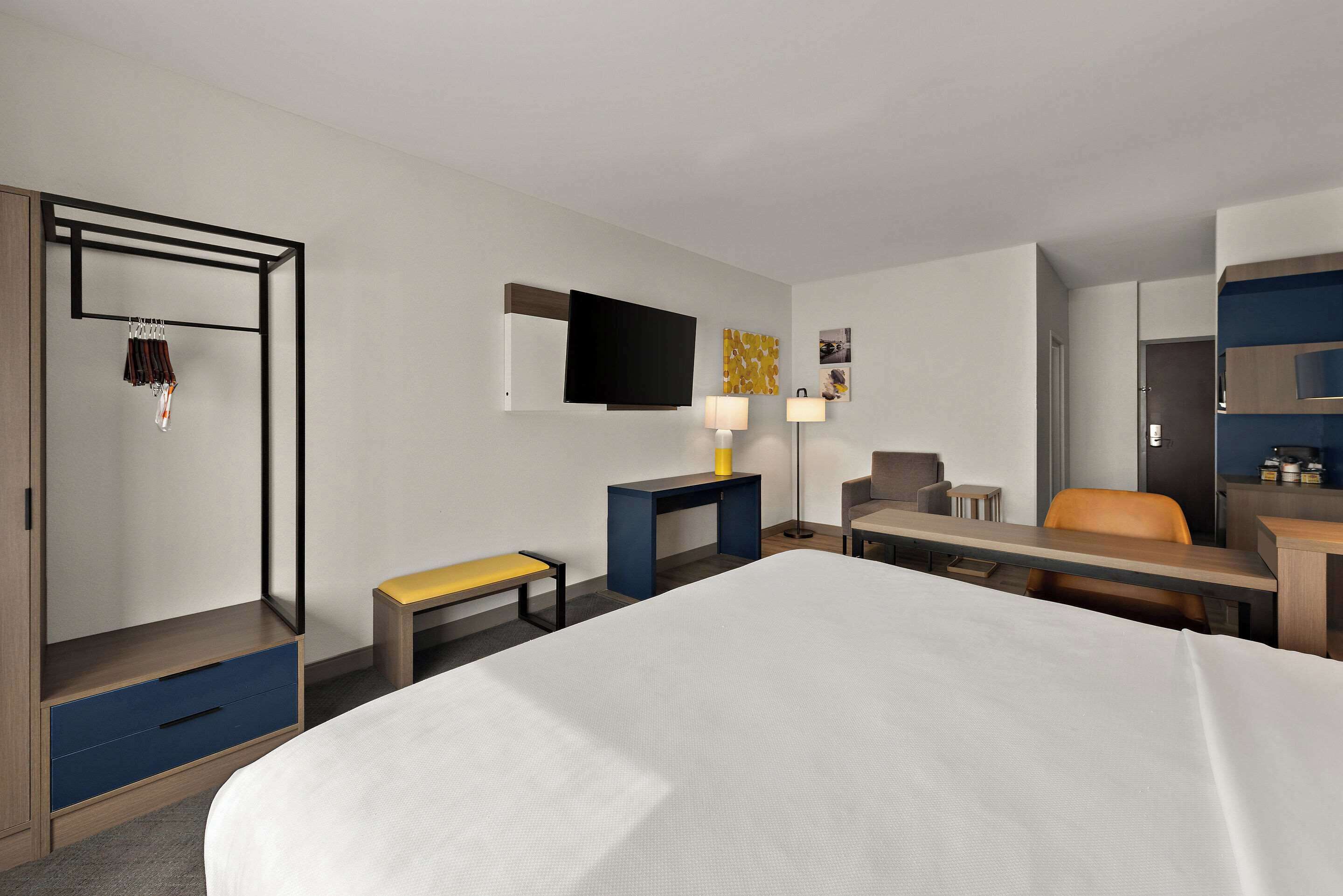 Ein Hotelzimmer mit einem großen Bett, einem Schreibtisch mit Stuhl, einem Fernseher, einem Kleiderschrank und einer Bank.