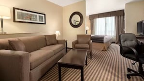A hotel room with a sofa, armchair, a small table, a bed, a TV, and a window with curtains. - University Hotel - Davis Area by IHG (Dixon)