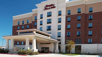 Un hotel Hampton Inn & Suites con fachada de ladrillo de dos pisos, un acceso cubierto con columnas y un cielo azul despejado al fondo.