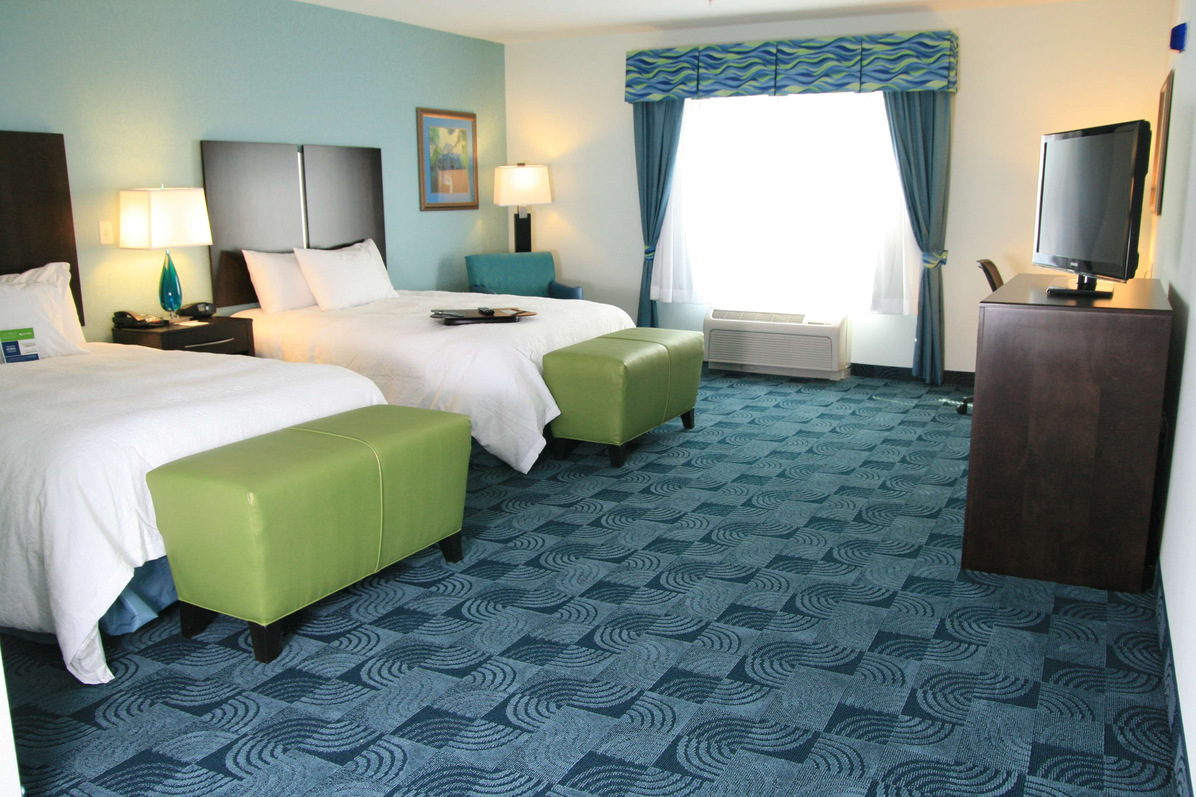 A hotel room with two beds, a desk with a TV, a desk lamp, a nightstand, a window with curtains, and a patterned carpet.