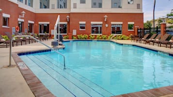 Una piscina con borde de ladrillo, sillas de descanso y un edificio con ventanas y un reloj.