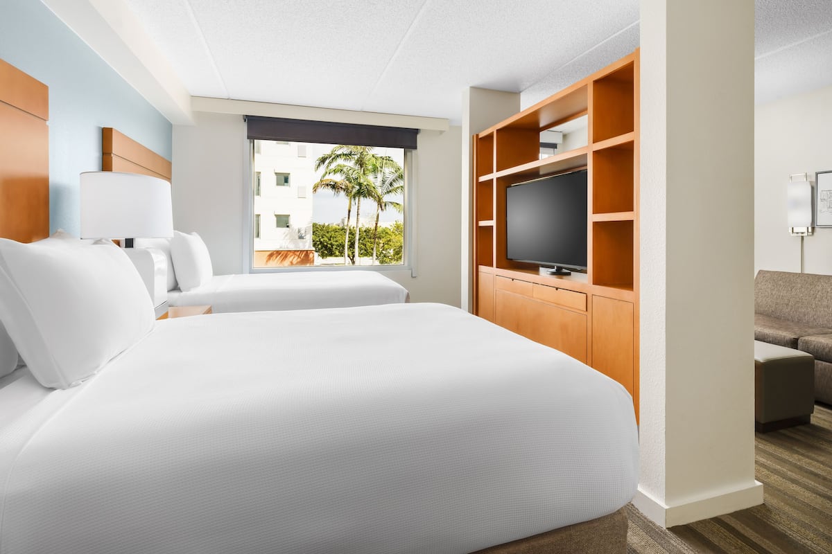 Une chambre d’hôtel avec un grand lit, un téléviseur posé sur un meuble en bois, une fenêtre donnant sur des palmiers et un canapé.