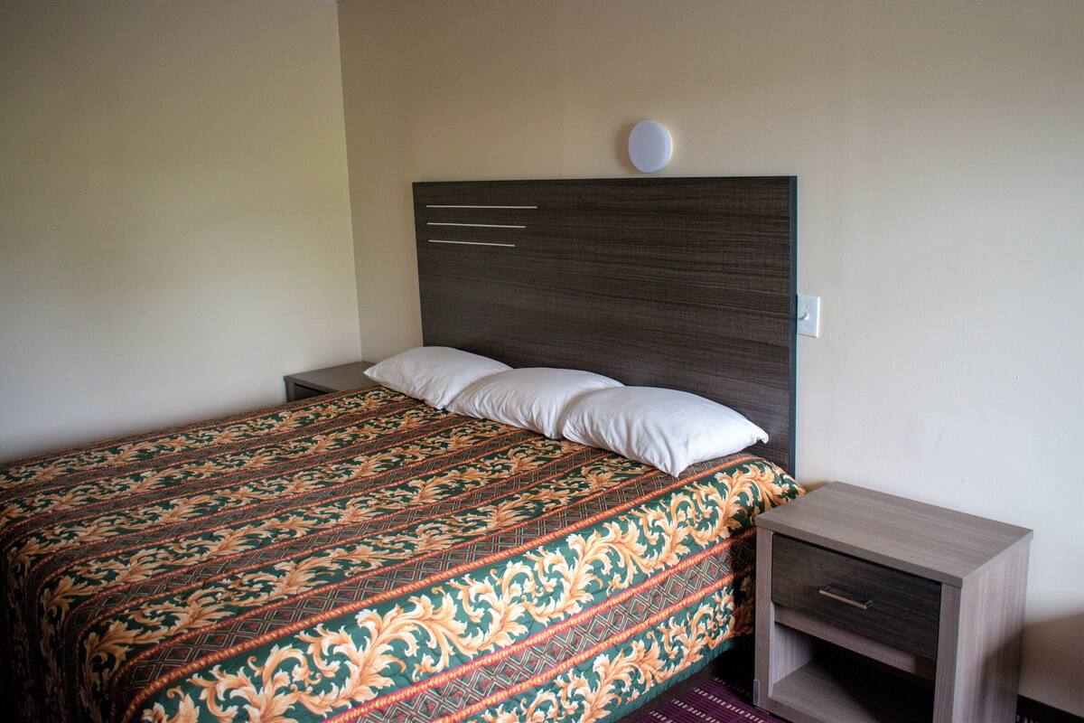 A neatly made bed with a patterned bedspread, a bedside table with a drawer, and a wall-mounted light fixture.
